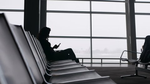 Young woman using smartphone in waiting room in airport,travels Mens come and stand, gaze out airport terminal window, silhouette view.