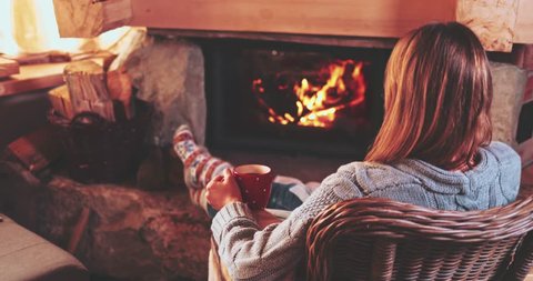Woman relaxes by warm fire with a cup of hot drink and warming up her feet in woollen socks. Feet in woollen socks by the Christmas fireplace. Cozy atmosphere. Winter and Christmas holidays concept. Arkivvideo