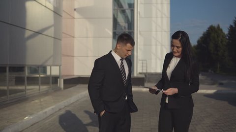 Business meeting. Young beautiful businesswoman talks and shows documents to young male employee outdoors against backdrop of business center