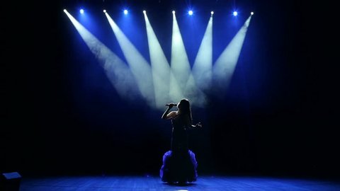 Silhouette of a beautiful girl on stage with concert lighting in a beautiful evening dress. The beautiful singer sings on stage in the dark.