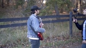 Using a gimbal, a teen filmmaker walks backwards filming a music video with a guitar player walking with a wood fence around a barnyard 
