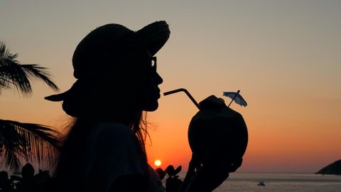 Silhouette of Young Mixed Race Hipster Girl Drinking Fresh Thai Coconut Water Coctail on the Beach Against Beautiful Sunset. 4K, Slowmotion. Phuket, Thailand.