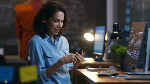 Beautiful Female Financier Uses Mobile Phone, Has Great News and Shows "Yes" Gesture, Her Personal Computer Showing Graphs and Statistics. She Smiles Charmingly.   
