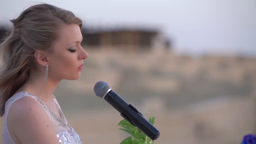 Elegant young blonde woman sings, plays romantic music on white digital piano in big arena or amphitheater. Girl in shiny evening dress performs on stage. Dubai Bab Al Shams Arena