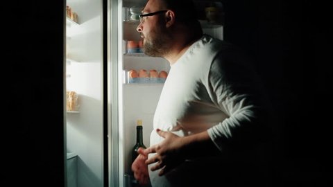 Fat man opens a refrigerator at night and is looking for something to eat, night hunger, gluttony concept