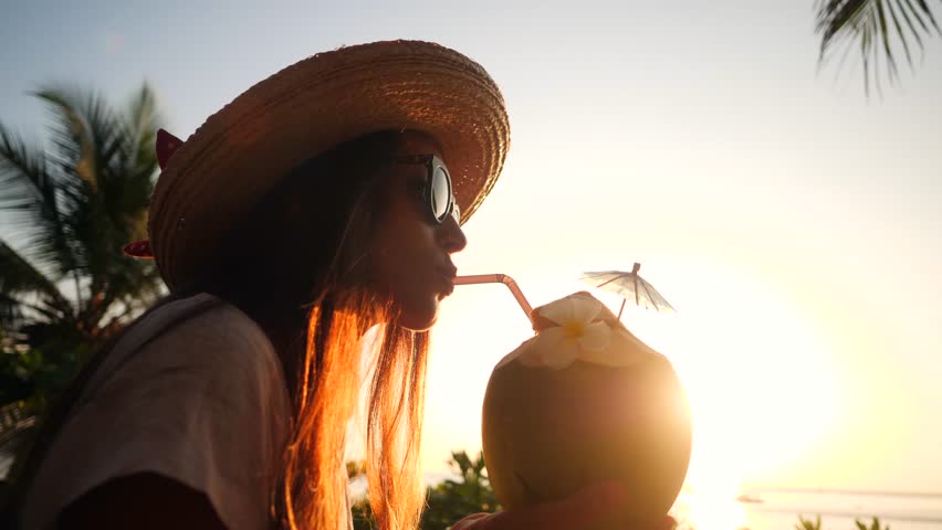 Attractive Young Tourist Hipster Girl Drinking Fresh Thai Coconut Water Cocktail at the Beach Against Beautiful Sunset. 4K, Slowmotion. Phuket, Thailand.