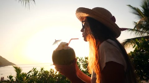 Attractive Young Mixed Race Tourist Girl Drinking Fresh Thai Coconut Water Coctail at Beach Against Beautiful Sunset. 4K, Slowmotion. Phuket, Thailand.