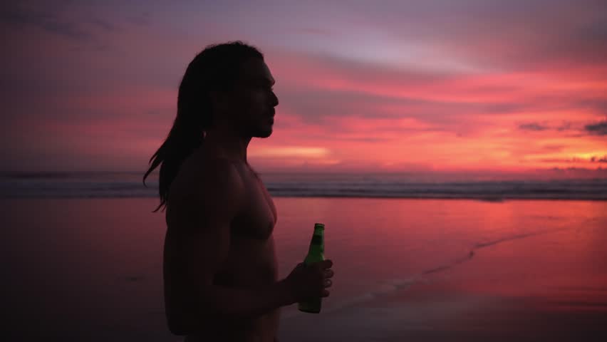 Young Man Drinking Beer From Glass Bottle At Beach Pink Sunset. Male Surfer With Dreadlocks And Alcohol Beverage At Gorgeous Summer Sea Or Ocean Water. Person Chilling With Lager At Seaside Shore Bank Royalty-Free Stock Footage #3378200417