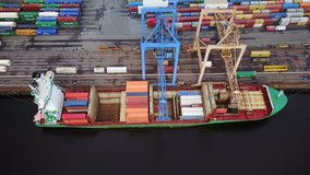 top down view of cargo being unloaded from a ship, docked at port. video descends downwards and tilts to reveal the containers being offloaded