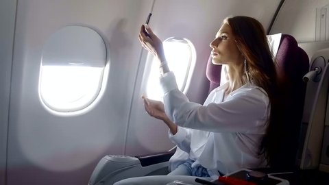 young pretty woman is applying cosmetic powder on her face, sitting on a seat in a modern airplane