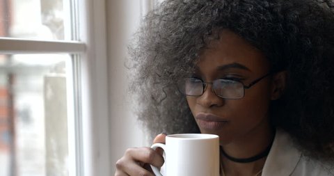 Young attractive afro-american woman in glasses is drinking tea while sitting on the window sill. Close-up portrait. 4k.