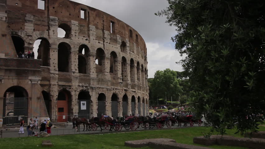 View of the Colosseum with tourists and horse carriages seen in front.