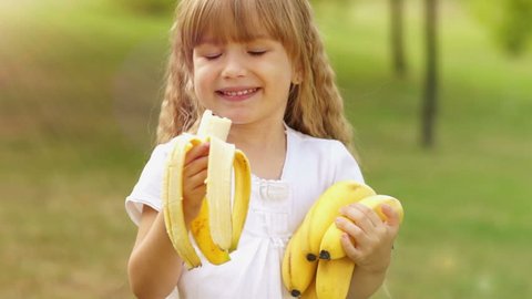 Sunny girl with bananas. The child looks at the camera. Happily eating fruit