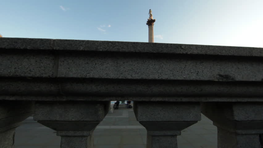 Camera move from behind a pillar structure revealing Trafalgar Square in London.