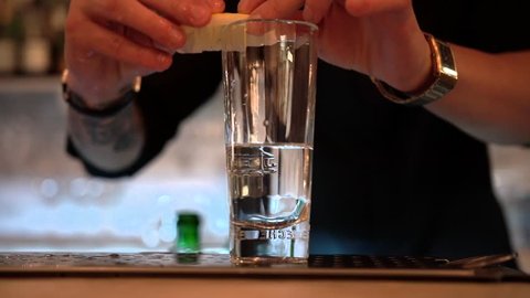 A bartender silhouette put a lemon slice on a transparent glass with alcohol drink on a bar background, close-up. Slow motion with blur bokeh lights.