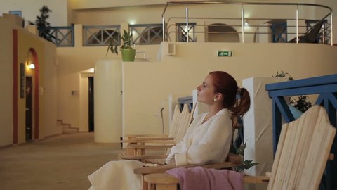 The portrait of woman who is sitting in the comfortable wooden armchair in luxury spa salon. The lady has red hair and wears white bathrobe while enjoying her relaxation and looks around. The room is
