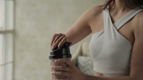 Attractive woman drinks protein cocktail is in apartment. Female opens plastic shaker, takes sip of wholesome beverage and smiles friendly, exposing teeth. Beautiful brunette dressed in trendy white