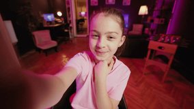 Kid films POV style video with smartphone in pink neon lit living room used as professional studio. Young online star captures footage with selfie cellphone camera, discussing about fun day at school