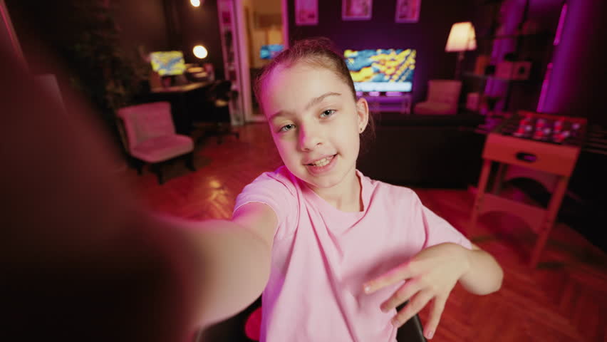 Kid films POV style video with smartphone in pink neon lit living room used as professional studio. Young online star captures footage with selfie cellphone camera, discussing about fun day at school Royalty-Free Stock Footage #3387605265
