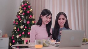 Asian female friends are watching a series through a laptop, chatting with their friends at the dining table in the festive atmosphere of Christmas at her home. Capture the cozy holiday vibes