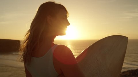 SLOW MOTION LENS FLARE: Lovely young surfer girl looking at golden sunset turns around and faces camera. Woman holding a surfboard, observing at sunset in Fuerteventura, Canary Islands turns around.