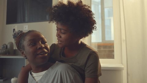 Medium close-up portrait shot of young single African American mother in health worker uniform posing in kitchen with 10-year-old son, who is hugging her, looking at camera and smilingの動画素材