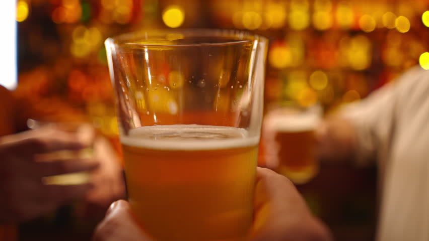 Close-up of a group of three friends standing at the bar talking and toast with glasses of beer and a cocktail in club bar