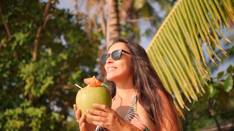 Attractive Happy Mixed Race Young Tourist Woman Drinking Fresh Thai Coconut Water Cocktail at Tropical Beach. 4K, Slowmotion. Phuket, Thailand.