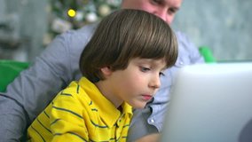 Father and son using laptop. Handsome father looking at computer with very cute son on a sofa at home on the background of Christmas tree. slow-motion. Father's Day. Christmas at home