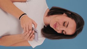 Vertical video, Beautiful smiling brunette young woman dressed in white top hold using mobile phone isolated on blue background in studio
