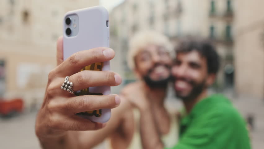 Close-up of hand, gay couple stands hugging and takes selfie on mobile phone, background blurre