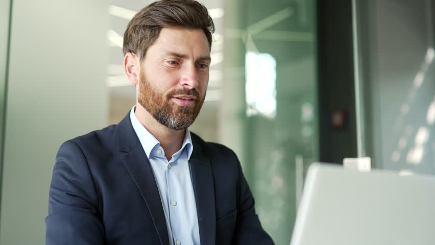 Smiling businessman in a formal suit typing on a laptop sitting at a workplace in business office. Handsome entrepreneur working on a project on computer, texting a client or chatting online. Close up