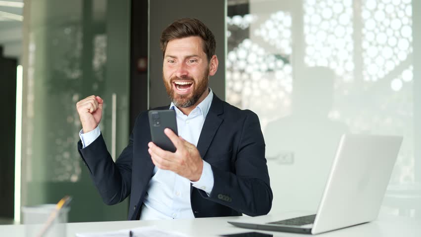 Excited happy businessman in formal suit received great news on smartphone sitting at workplace in business office. A joyful entrepreneur reads a pleasant message on a mobile phone, celebrates success | Shutterstock HD Video #3389379051