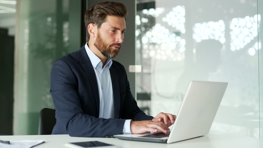 Upset businessman suffering from toothache working on laptop sitting at workplace at desk in modern office. Entrepreneur holds his hand on his cheek, massaging the sore spot. He needs to see a dentist Royalty-Free Stock Footage #3389395689