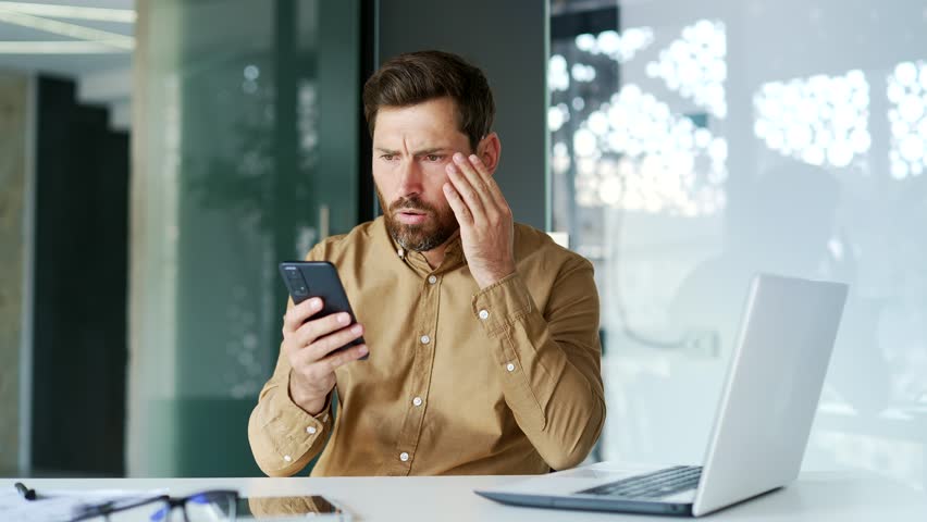 Frustrated stressed manager reads bad news on a smartphone while sitting at workplace in business office. Disappointed entrepreneur or worker is upset reviewing negative information on mobile phone | Shutterstock HD Video #3389434239