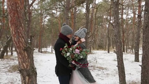 Newlyweds groom and bride hug kiss and warm each other in snowy pine forest during snowfall in slow motion. . Young couple in winter wood. Woman with bouquet. Valentine's Day and Christmas concept.