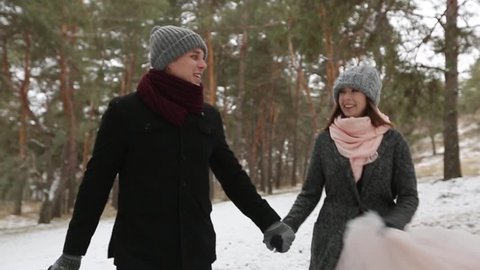 Outdoor winter shot of young wedding couple running and having fun holding hands in snow weather pine forest during snowfall. Snowy engagement ceremony.