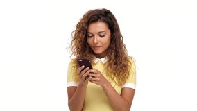 Happy surprised curly brunette woman in dress writing message on smartphone over white background