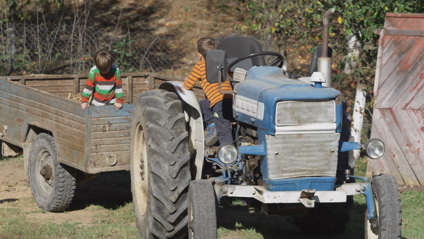 Cute little kids playing with farm tractor, big brother driving the steering wheel while little sibling stands in the trailer tow, fun at countryside
