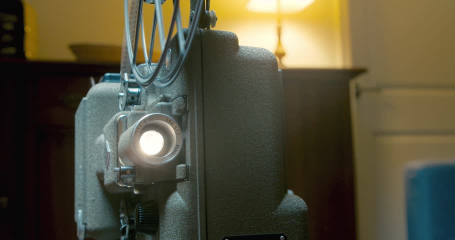 Vintage 8mm film projector spins a reel with a movie in the evening in a home interior. An old film projector with a film on the background of an old lamp and old furniture.