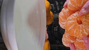 vertical video Place peeled tangerines on a white plate and divide them into slices. Female hands prepare citrus fruits for tangerine juice.