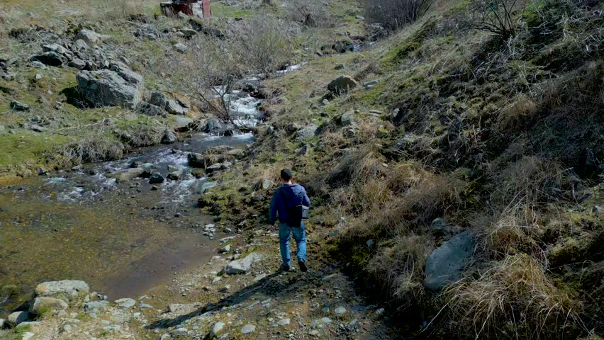 Aerial view. A man in blue clothes is walking along a muddy stony country road, crossing a mountain river on stones. Around the gray mountains without grass. Aerial view, drone footage. Royalty-Free Stock Footage #3390036767