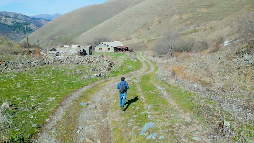 Aerial view. A man in blue clothes walking along a muddy stony country road along old ruined houses. All around are gray mountains without grass. Aerial view, drone footage. Royalty-Free Stock Footage #3390048421