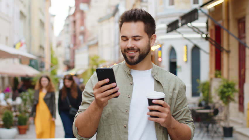 Happy smiling man use smartphone typing text messages browsing internet social media web app working chatting online enjoying morning to-go coffee hot drink outdoor. Guy tourist walking in city street Royalty-Free Stock Footage #3390203969