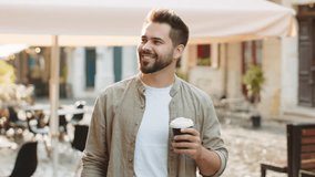 Happy bearded young man enjoying morning coffee hot drink and smiling outdoors. Relaxing, taking a break. Guy walking in urban city sunshine street, drinking coffee to go. Town lifestyles outside