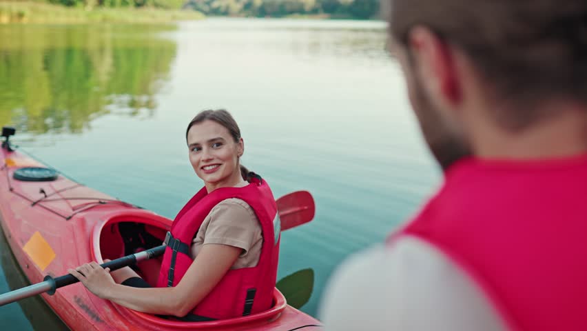Rear view on blurred man speaking with his lovely woman in kayak. Beautiful female with safety vest holding oar while sitting in red boat on lake. Positively smiling. Kayaking activity. | Shutterstock HD Video #3390453305