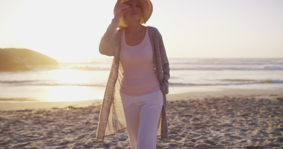Senior woman, happy and walking on beach with health wellness and positive on weekend vacation. Mature person, relax and summer hat for adventure by ocean, travel and calm joy on retirement by sunset Royalty-Free Stock Footage #3390543807