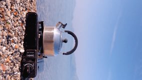 View of metal kettle boiling on portable gas stove, against background of calm lake and mountains at dawn, traveling and relaxing in nature. Vertical video