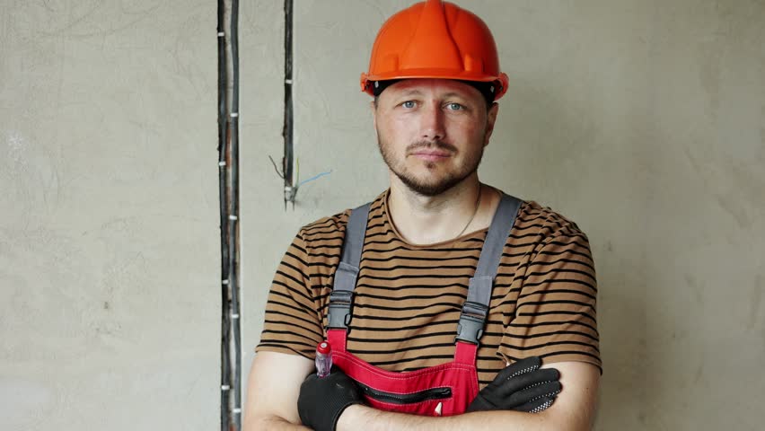 Renovation apartment. Portrait of mature confident male foreman constructor repairman with crossed hands in safety hard hat and overalls looks at camera, holds screwdriver. Empty concrete walls Royalty-Free Stock Footage #3390723701