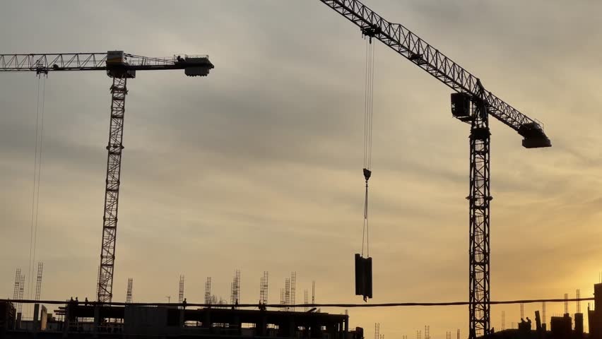 Mooving silhouette of a construction crane at an urban building site. Workers are visible against the fading light, showcasing the scale and ongoing progress of the development.